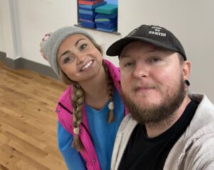 A man with a black hat and a beard smiling, with a young woman with a grey bobble hat smiling.